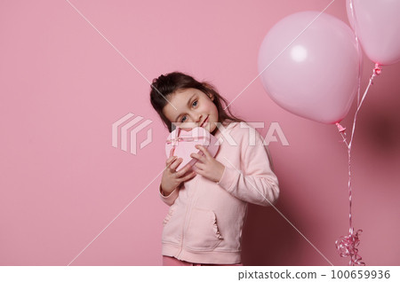 Beautiful Caucasian little girl, enjoying festive occasion event, holding a cute heart shaped gift box, smiling, isolated on pink color background with pink pastel air balloons. Mother's Day concept Beautiful Caucasian little girl, enjoying festive occasion event, holding a cute heart shaped gift box, smiling, isolated on pink color background with pink pastel air balloons. Mother's Day concept 100659936