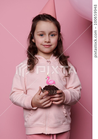 Vertical advertising studio shot on pink color background, lovely little child girl in pink party hat with a birthday cake in her hands, cutely smiling looking at camera. Happy Childhood concept 100659948