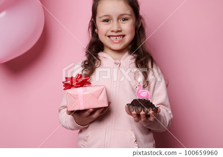 Focus on a gift box with red bow and a cupcake with candle, in the hands of a blurred charming little child girl, enjoying her birthday party over isolated pink color background. Happy childhood 100659960