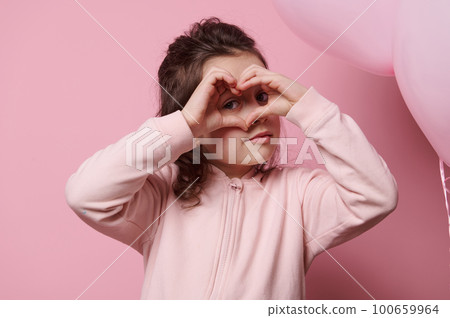 Beautiful Caucasian little child girl looking through a heart made from her fingers, gesturing with hand sign, showing you her affection, sympathy and love, isolated over pink colored background 100659964