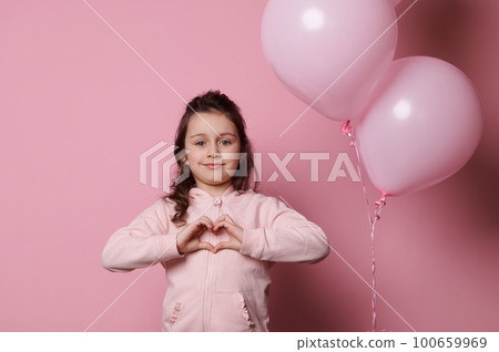 Adorable Caucasian child girl in casual pink clothes, shows a heart made with her fingers, smiles at camera, isolated over pink colored background with inflatable helium pink pastel balloons Adorable Caucasian child girl in casual pink clothes, shows a heart made with her fingers, smiles at camera, isolated over pink colored background with inflatable helium pink pastel balloons 100659969