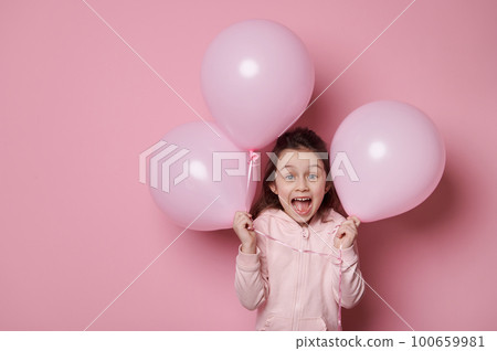 Amazed little child girl in pink hoodie, holding pink pastel balloons, expressing surprise and amazement, posing with open mouth on isolated pink color background with copy advertising space for text 100659981