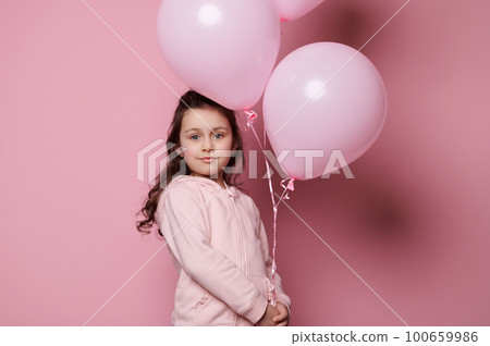 Adorable little child girl holding a bunch with inflatable pink pastel balloons, isolated on pink color background, confidently looking at camera. Emotions. Kids. International Children's day concept Adorable little child girl holding a bunch with inflatable pink pastel balloons, isolated on pink color background, confidently looking at camera. Emotions. Kids. International Children's day concept 100659986