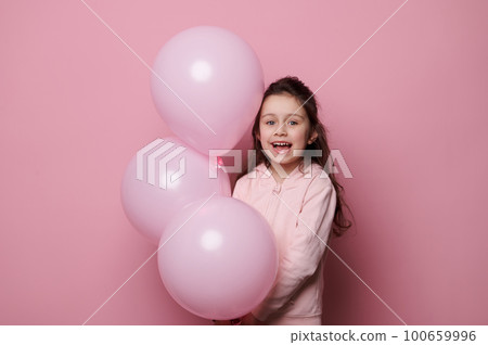 Caucasian beautiful little child, birthday girl in pink clothes, smiling looking at camera, posing with pink pastel helium balloons, isolated over pink color background with copy advertising space 100659996