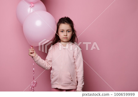 Caucasian elementary age child girl with a bunch of pink pastel balloons, looking at camera, isolated over pink color background with copy advertising space. Birthday party. Celebrating festive events 100659999