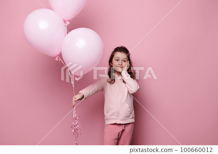 Adorable 5-6 years Caucasian child, lovely little girl in pink clothes, holding a bunch of pink pastel helium balloons, cutely smiles, looking dreamily aside, isolated on pink color background 100660012