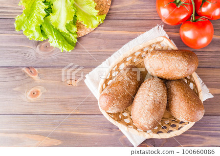 Fresh rye buns in a basket, salad and tomato on a wooden table. Top view. Copy space 100660086