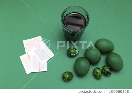 Eggs painting dye, colorant in paper bag, package. Glass with Colored water, Green Easter hen, quail eggs on blue green background. Top view, flatly horizontal plane. High quality photo. Eggs painting dye, colorant in paper bag, package. Glass with Colored water, Green Easter hen, quail eggs on blue green background. Top view, flatly horizontal plane. High quality photo. 100660238