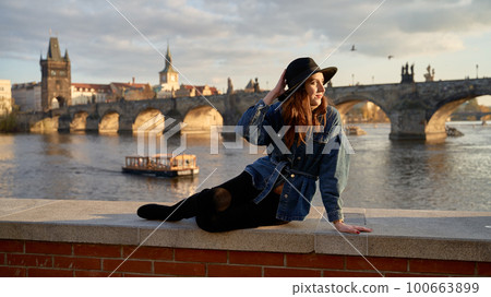 Stylish beautiful young woman wearing black hat sitting on Vltava river shore in Prague with Charles Bridge on background. Elegant retro lady fine art portrait. 100663899