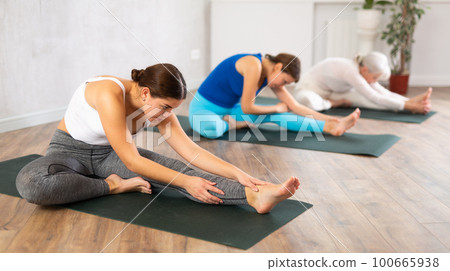 Yoga class of relaxed sports women performing Head to Knee Forward Bend Pose, or Janu Sirsasana after practicing together in gym Yoga class of relaxed sports women performing Head to Knee Forward Bend Pose, or Janu Sirsasana after practicing together in gym 100665938