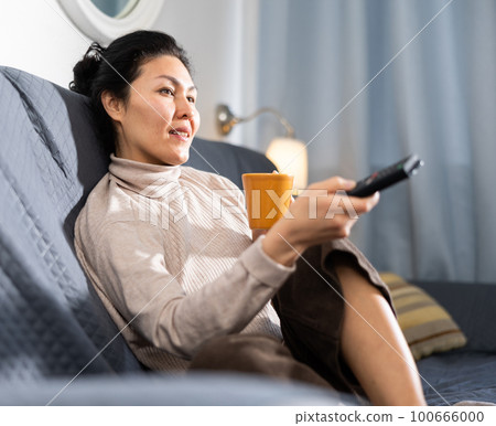 Woman with cup of tea sitting on sofa and watching television Woman with cup of tea sitting on sofa and watching television 100666000