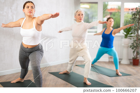 Diligent women practicing warrior pose of yoga in light fitness room Diligent women practicing warrior pose of yoga in light fitness room 100666207