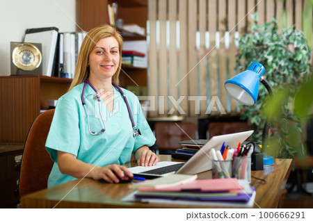 Positive woman doctor working in cabinet on laptop Positive woman doctor working in cabinet on laptop 100666291