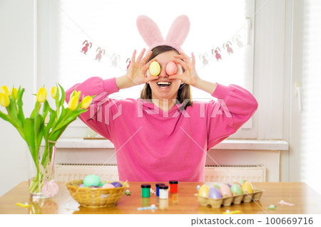 Happy beautiful young woman smiling wearing rabbit ears holding Easter eggs for her eyes. 100669716
