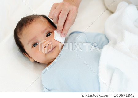 mother cleaning and wiping newborn baby face with cotton pad on bed 100671842