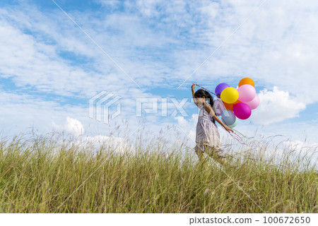 Cheerful cute girl holding balloons running on green meadow white cloud and blue sky with happiness. Hands holding vibrant air balloons play on birthday party happy times summer on sunlight outdoor 100672650