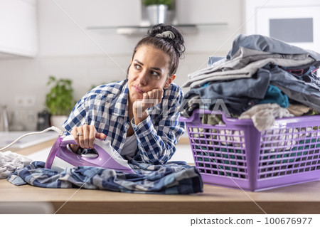 Woman is daydreaming while ironing next to an overfilled big basket full of clothes 100676977