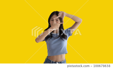Playful young asian woman in good mood posing for photo or photo frame with fingers on yellow background,  Use your fingers to act like a photograph, Mark as if taking a photo. 100678638