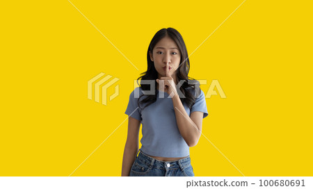 Asian woman doing silent gesture with finger, Noiseless symbol, Noisy mall, Hide a secret, Do not speak. shut your mouth, Noiseless signal transmission, Isolated on yellow background. 100680691