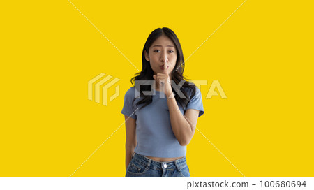 Asian woman doing silent gesture with finger, Noiseless symbol, Noisy mall, Hide a secret, Do not speak. shut your mouth, Noiseless signal transmission, Isolated on yellow background. 100680694