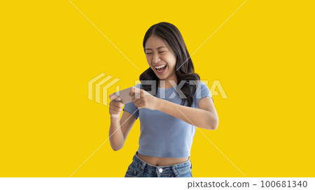 Asian woman playing games on mobile phone and wearing headphones having fun, Play games on the sofa in the living room on weekends, Resting at home, Comfort zone, Touch screen mobile phone. 100681340