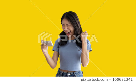 Asian woman playing games on mobile phone and wearing headphones having fun, Play games on the sofa in the living room on weekends, Resting at home, Comfort zone, Touch screen mobile phone. 100681341