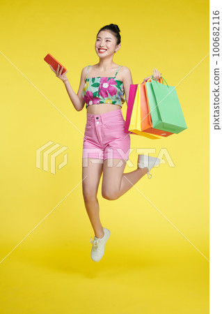 Full length of happy young attractive shopaholic woman raising shopping bags and holding mobile phone Full length of happy young attractive shopaholic woman raising shopping bags and holding mobile phone 100682116