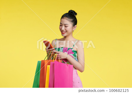 Portrait of a cheerful young girl in summer clothes holding mobile phone with shopping bags isolated over yellow background 100682125