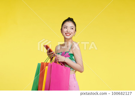 Young excited cheerful woman holding shopping bags isolated over yellow background using mobile phone looking aside. Young excited cheerful woman holding shopping bags isolated over yellow background using mobile phone looking aside. 100682127