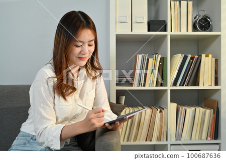 Pretty young woman sitting on couch at home, using digital tablet for distant studying or online communication 100687636