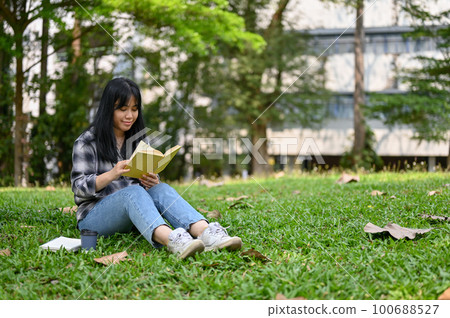 Pretty Asian female college student sits on grass and reads a book in the campus park. Pretty Asian female college student sits on grass and reads a book in the campus park. 100688527