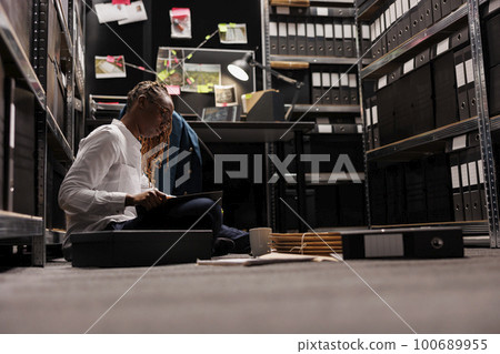 Lawyer preparing for case and reading detective report while sitting on floor. African american investigator analyzing csi expertise and evidence while working overtime at night 100689955
