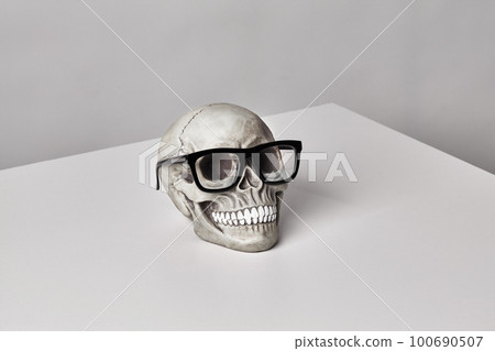 Realistic model of a human skull with teeth in a black-framed classic glasses on a light table, white background. Halloween horror concept. 100690507