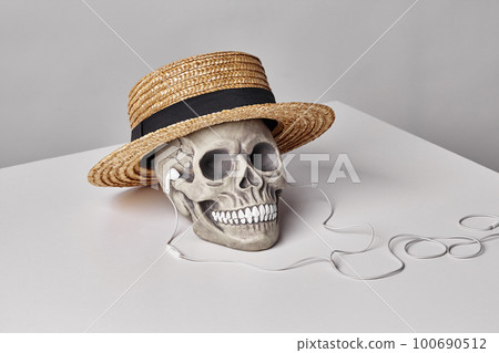 Realistic model of a human skull with teeth in a yellow straw hat and headphones on a light table, white background. Halloween horror concept. 100690512