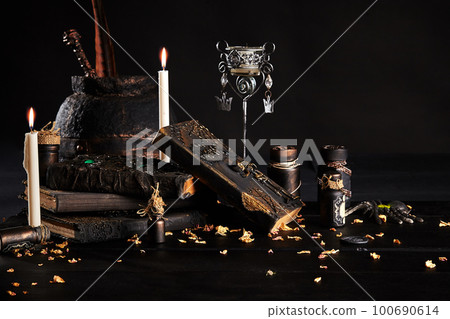 Books with spells, old pot, jars of potion are on a wooden dark table. Petals of dried roses are scattered nearby. Black background. Close-up shot. 100690614