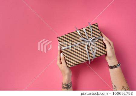 Hands of a woman with tattoos and watch holding striped brown gift box tied with silver ribbon on a pink background. Close-up, copy space, top view. 100691798