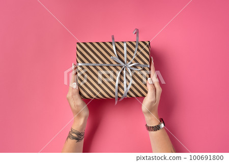 Hands of a woman with tattoos and watch holding striped brown gift box tied with silver ribbon on a pink background. Close-up, copy space, top view. 100691800