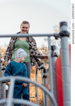 A boy, person with down syndrome walks in the park with his mother, going down the children's slide A boy, person with down syndrome walks in the park with his mother, going down the children's slide 100691867