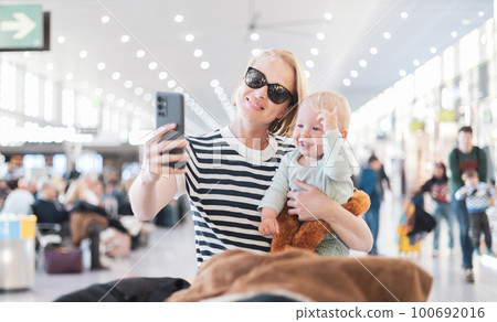 Mother taking selfie with mobile phone, while traveling with child, holding his infant baby boy at airport, waiting to board a plane. Travel with kids concept 100692016