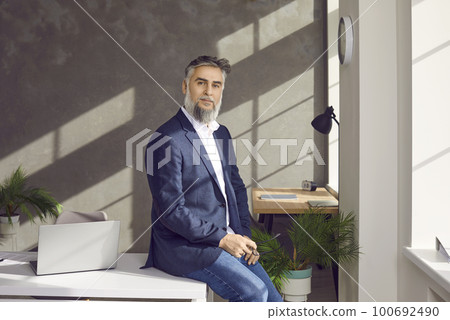 Mature business man sitting on the table of his workplace at office, looking confident at camera. 100692490