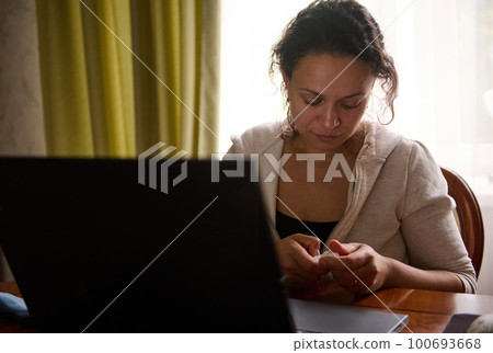 Multi-ethnic young woman reads instructions for the use of eye drops from dryness while working on laptop. Treating eyeball irritation or inflammation. Irritated eye, optical symptoms. Ophthalmology 100693668