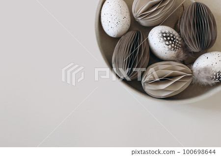Moody spring still life scene. Easter composition. Bowl of folded paper ornaments, hen eggs decorated by guineafowl bird feather. Beige table background. Neutral festive flat lay, top view. Web banner 100698644