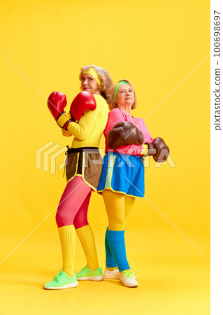 Active lifestyle. Elderly sportive women in colorful uniform posing in boxing gloves against yellow studio background. Concept of sport and health 100698697