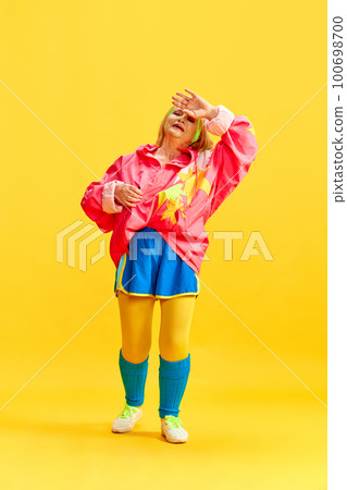 Wiping sweat after workout. Elderly sportive woman in colorful uniform training, posing against yellow studio background. Concept of sport and health 100698700