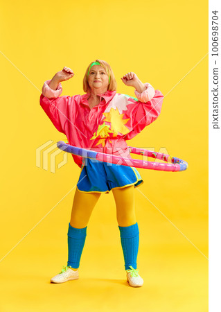 Elderly, cheerful, sportive woman in colorful uniform training, twisting hoop against yellow studio background. Concept of sport and health 100698704