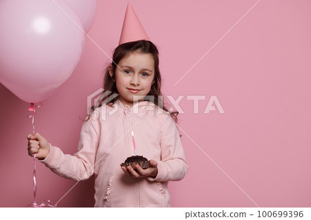 Caucasian 5 years old child, lovely birthday girl in pink party hat, smiling at camera, holding a bunch of inflatable pink balloons and a festive cake with candle, isolated on pink colored background Caucasian 5 years old child, lovely birthday girl in pink party hat, smiling at camera, holding a bunch of inflatable pink balloons and a festive cake with candle, isolated on pink colored background 100699396