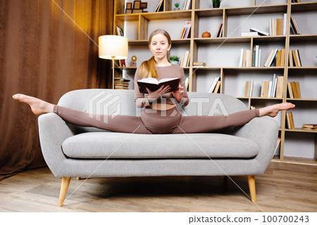 Young beautiful blonde woman in brown leggings and top sitting on a twine at home on the couch. The girl is reading a book sitting on the couch in an unusual position 100700243