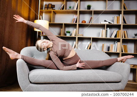 A cute sporty girl sits on the couch in a splits pose and stretches her hand to her leg A cute sporty girl sits on the couch in a splits pose and stretches her hand to her leg 100700244