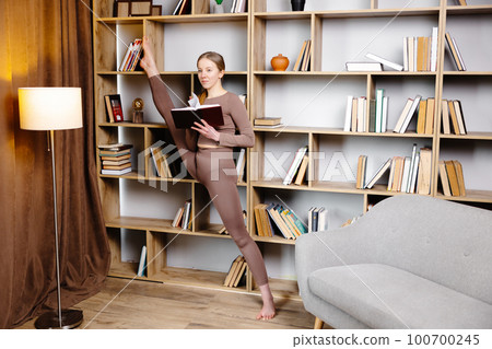 Young woman in sportswear stretching legs, doing splits standing in library and reading book. Female in daily workout make yoga training at home 100700245