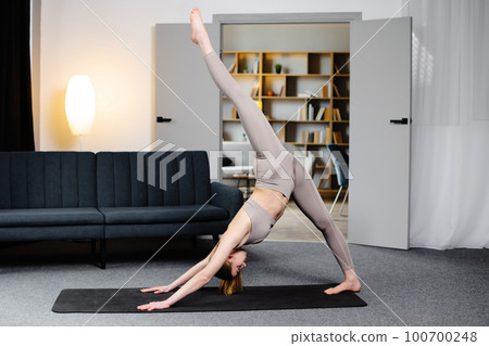 Side view of sporty girl in leggings and top standing in three-legged down dog pose in living room. Caucasian barefoot lady practicing yoga, focused on exercising along at home. Concept of yoga. 100700248